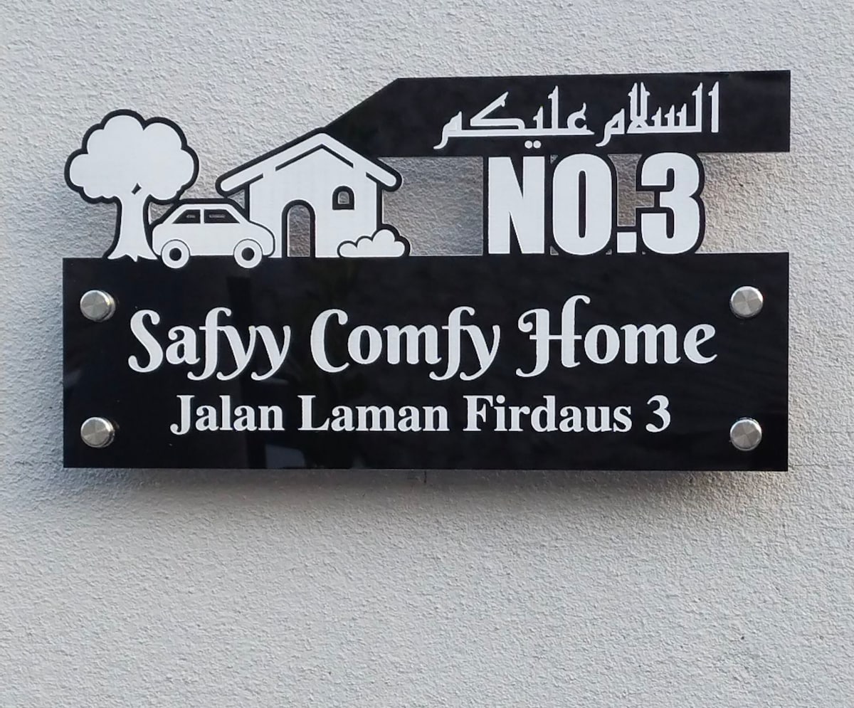 A wall-mounted sign displays the name 'Safyy Comfy Home' along with the address 'Jalan Laman Firdaus 3'. The design includes a tree and a house graphic, with a vehicle included for added context. The sign features a modern black and white color scheme.