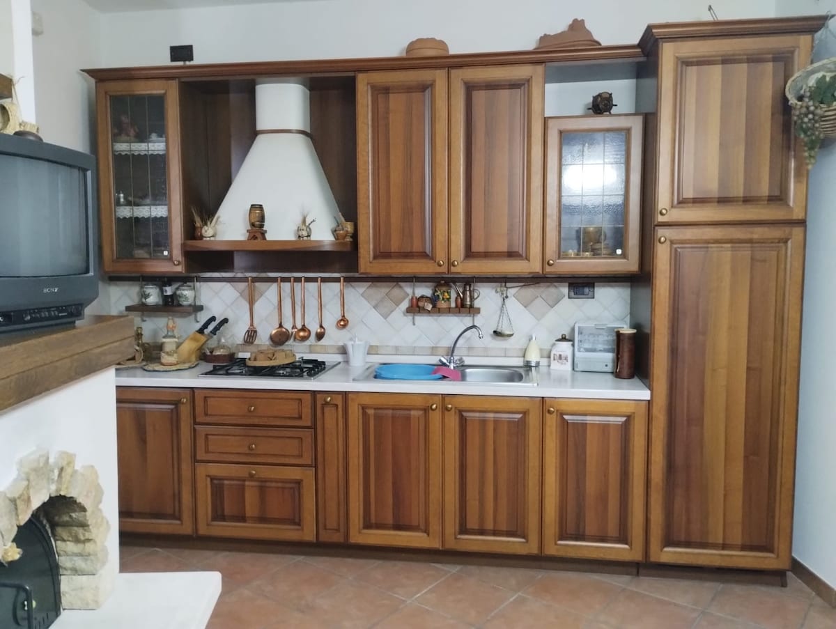 A wooden kitchen is featured with a range of cabinets and countertops. A traditional-style stove and hood are visible, accompanied by hanging cooking utensils. Additional appliances and decorative items can be seen on the countertops, complemented by a tile backsplash.
