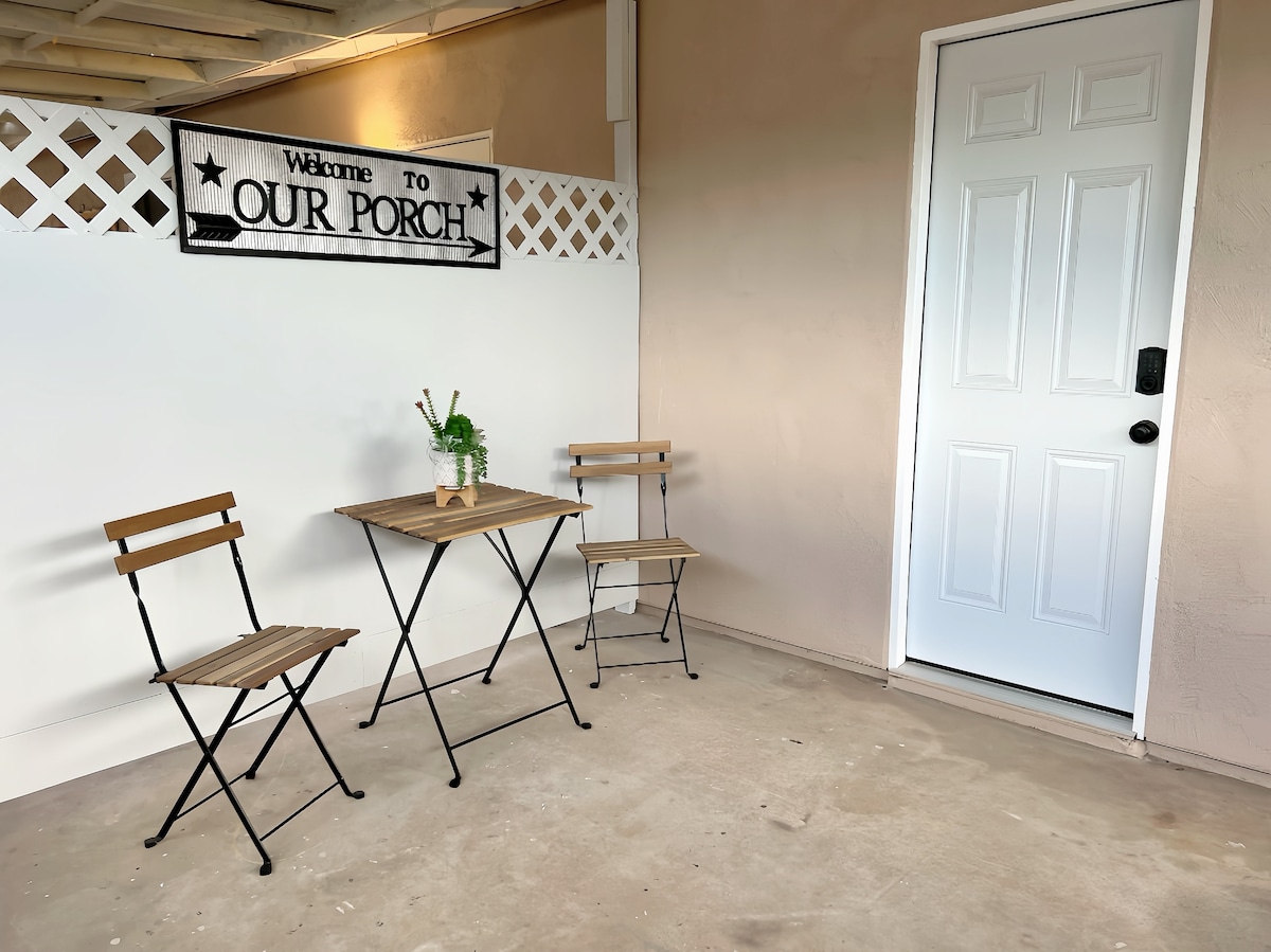 A small outdoor seating area features a round table flanked by two foldable wooden chairs. A decorative sign reading 'Welcome to Our Porch' adorns the wall above. The entrance door is situated on the right, contrasting against the light-colored wall.