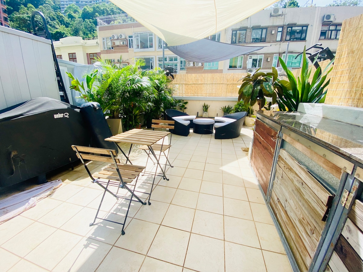 A spacious rooftop terrace is equipped with a Weber BBQ and lounge seating. Potted plants enhance the greenery of the space. Two foldable chairs and a small table are placed on the tiled surface, providing an area for relaxation or outdoor dining.