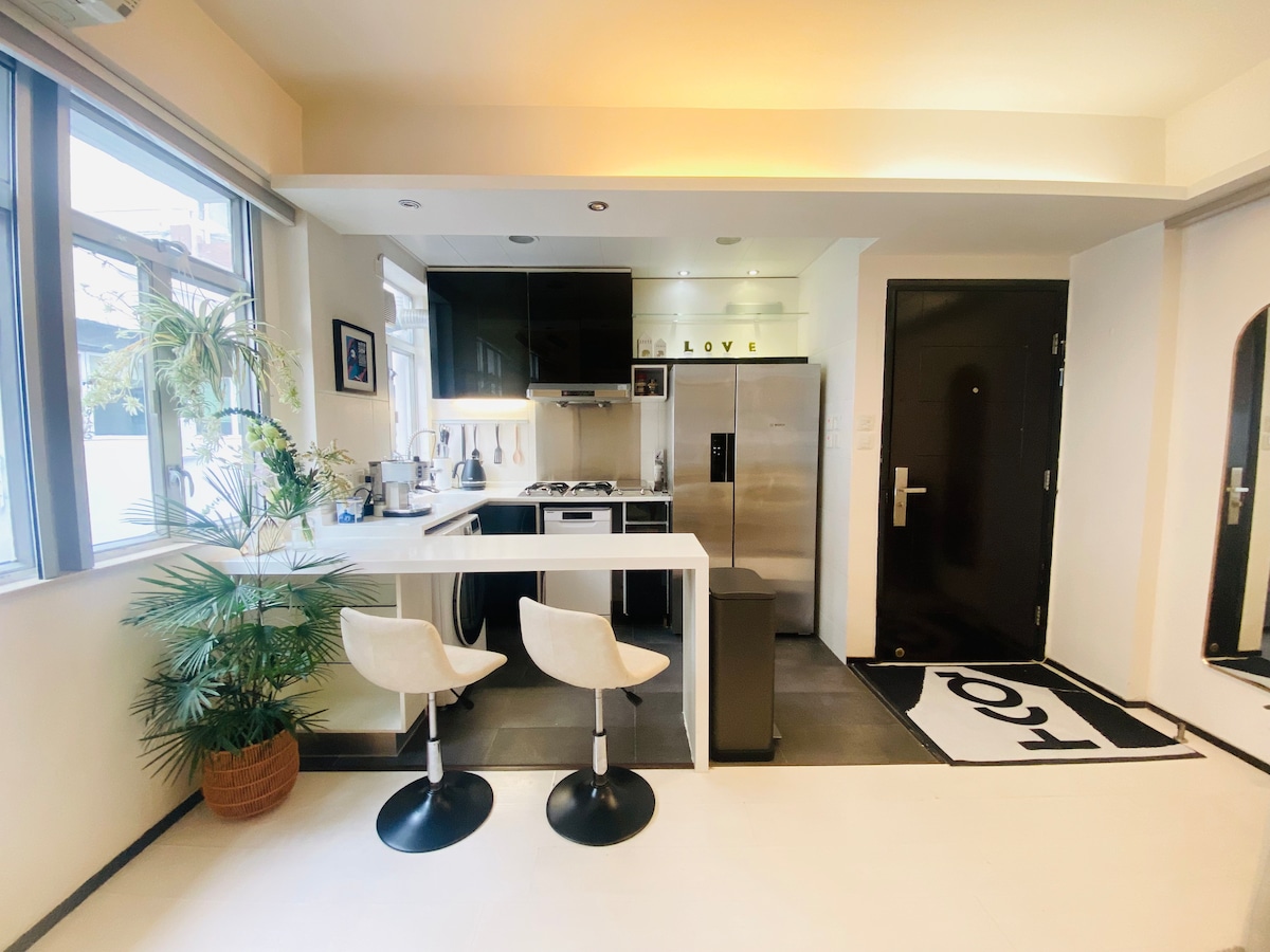 A modern kitchen area is seen, featuring a white bar table with two stools. Black cabinetry and stainless-steel appliances complement the space. A large window allows natural light to brighten the area, while a potted plant adds a touch of greenery.