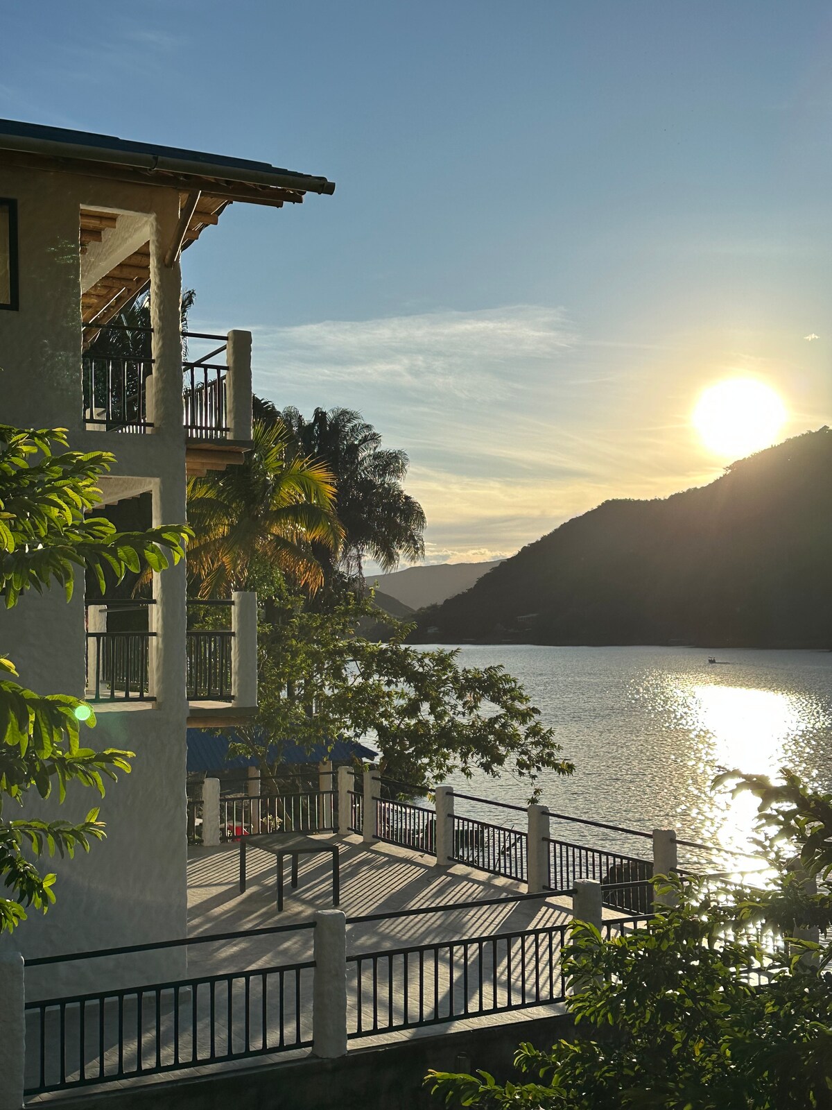 A view of the house's balcony overlooks a serene lake at sunset. The sun reflects on the water's surface, while lush greenery surrounds the area. The mountains in the background create a tranquil backdrop, enhancing the natural beauty of the setting.