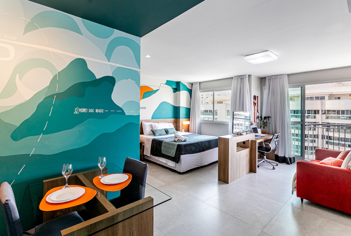 A modern studio space is highlighted by a large mural depicting mountains. The area features a spacious bed, a work desk with an ergonomic chair, and a cozy seating arrangement. Large windows provide natural light and views of the surrounding city.