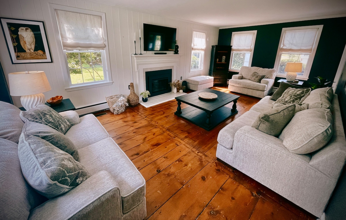 A spacious living room features large windows allowing natural light, a cozy arrangement of light gray sofas and cushions, and a central dark coffee table. A television is mounted above a white fireplace, complemented by a decorative owl artwork on the wall.