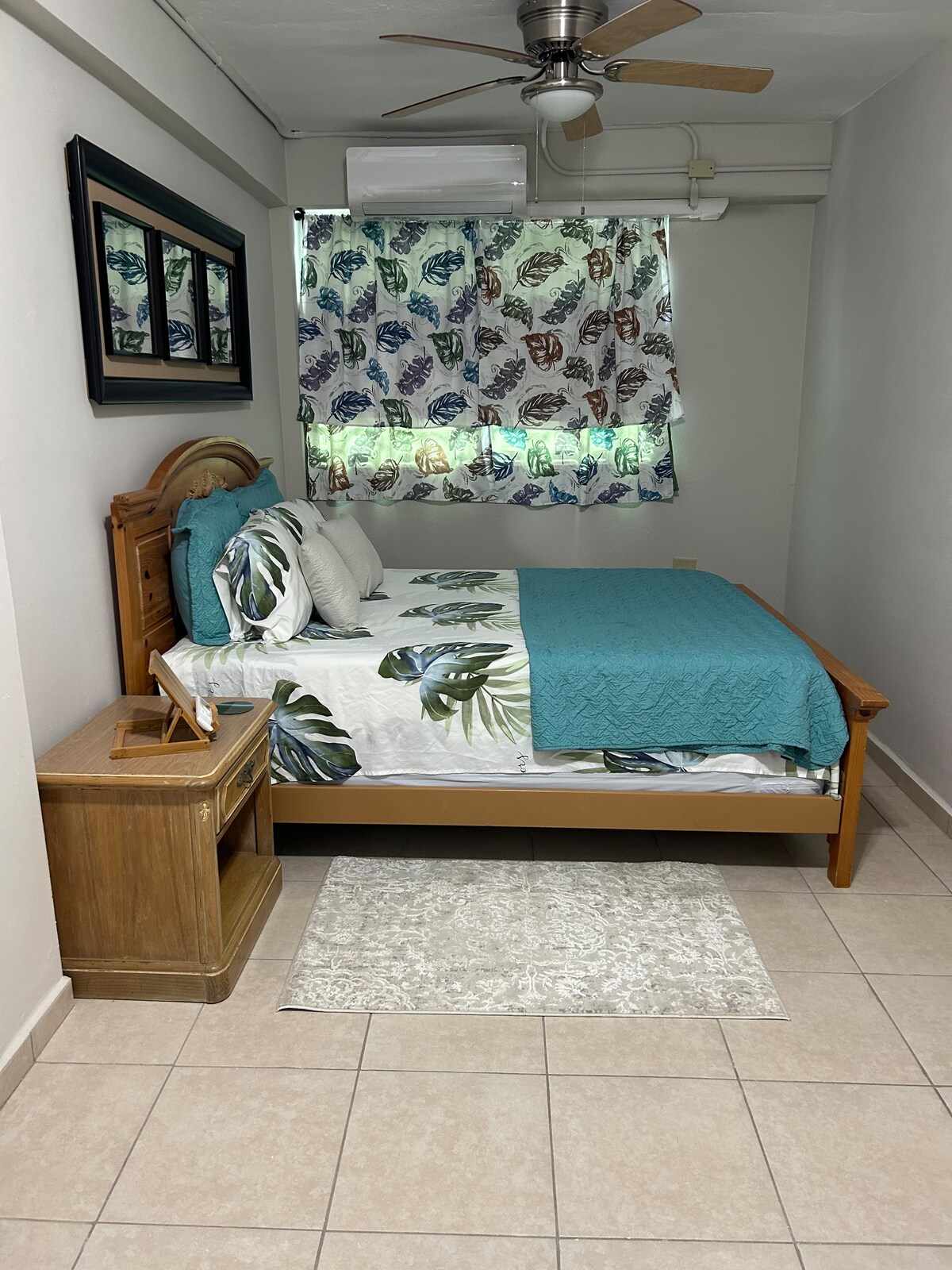 A cozy sleeping area features a wooden bed adorned with a turquoise and leaf-patterned comforter. Complementary green and white curtains allow natural light to filter in. A bedside table holds a small decorative item, with a light-colored rug positioned beside the bed on tiled flooring.