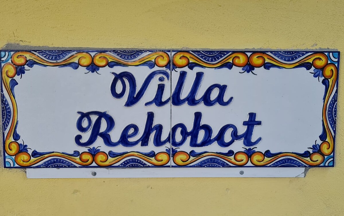 A ceramic tile sign displays the name 'Villa Rehobot' in blue script against a yellow background, framed by decorative orange and blue motifs. The sign is affixed to a textured wall, providing a welcoming entrance to the property.