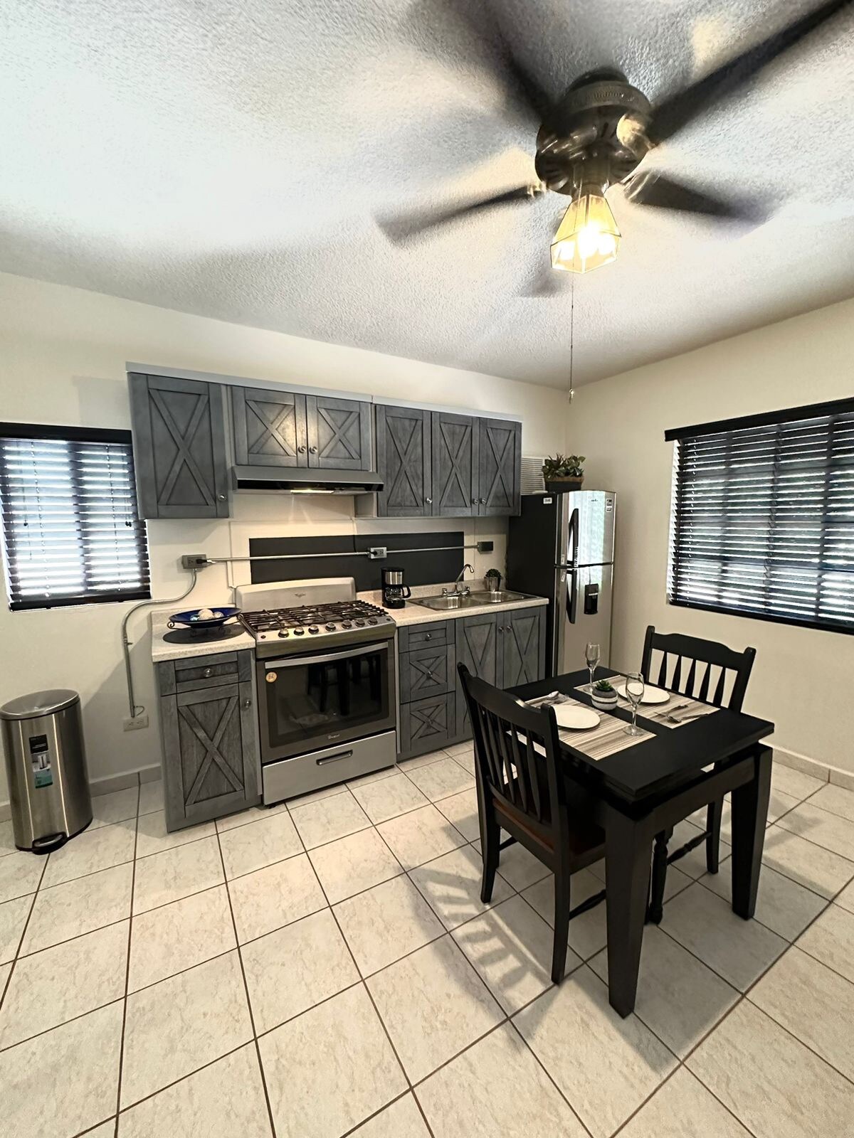 A modern kitchen is presented with gray cabinetry and matching appliances. A dining table for two is set against a wall, accompanied by two chairs. Blinds cover the windows, allowing controlled light entry, while a ceiling fan adds airflow to the space.