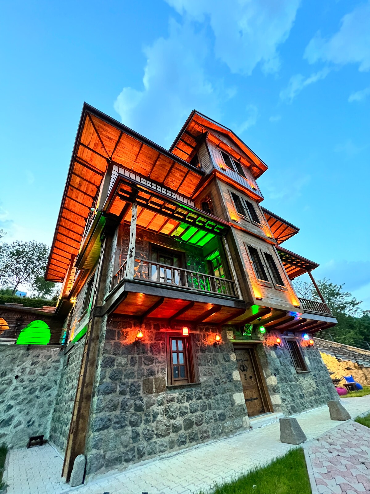A historic building is showcased from an exterior angle, featuring layered balconies and decorative wooden elements. The stone façade is enhanced by colorful lighting, creating a vibrant atmosphere. A spacious stone pathway leads to the entrance, surrounded by greenery in the background.