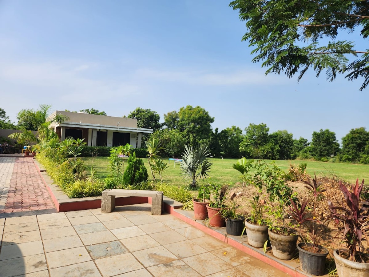 A spacious outdoor area is framed by a variety of potted plants. The scene includes a well-manicured lawn and a single-story building in the background, surrounded by trees. A pathway lined with red bricks leads to a stone bench, inviting relaxation in the serene environment.