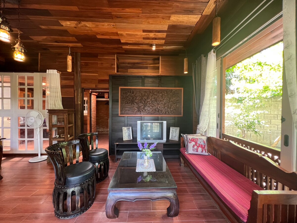 A wooden living area features dark wood paneling and flooring. A glass coffee table sits atop the tiles. Seating includes two carved chairs and a wooden bench adorned with vibrant cushions. Natural light filters in through large windows, enhancing the warm ambiance.
