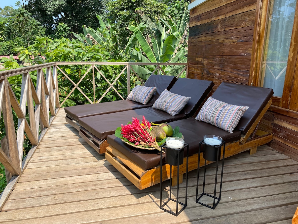 A wooden deck features two lounge chairs with brown cushions and striped pillows, surrounded by lush greenery. Small side tables with decorative candles and tropical fruits are positioned nearby, creating a tranquil setting for relaxation.