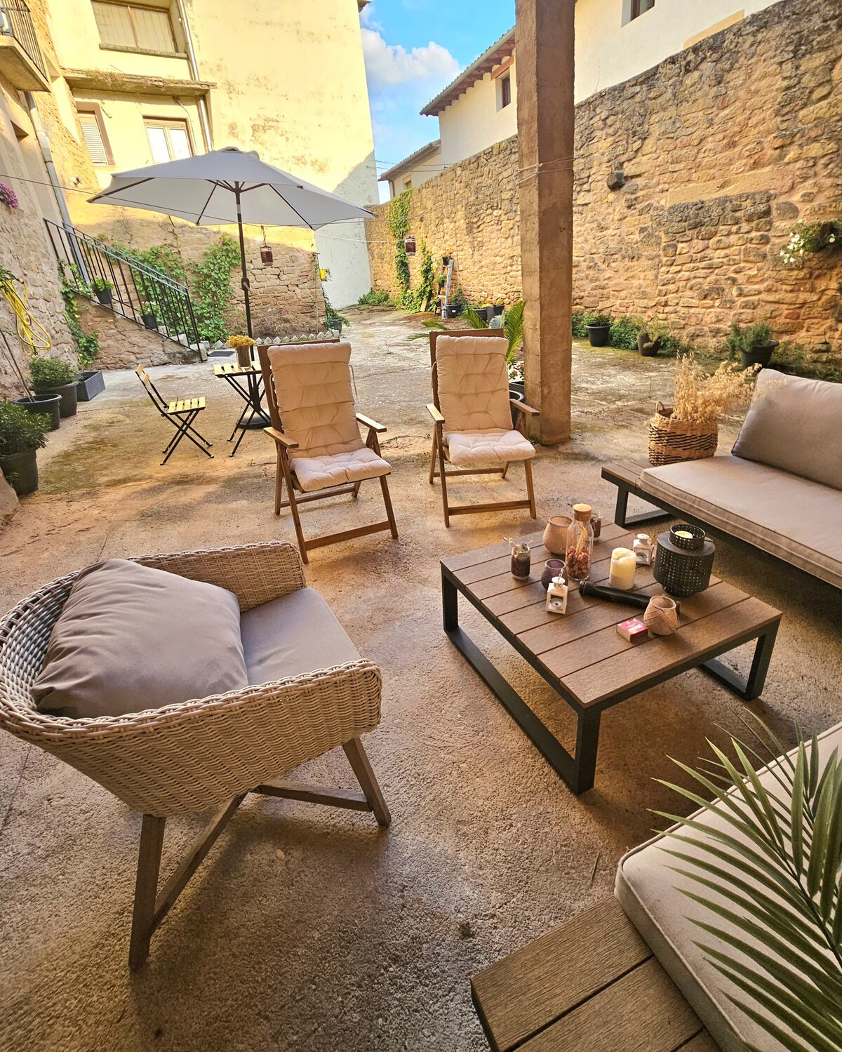 A private patio features seating arrangements with two lounge chairs and a comfortable sofa, accompanied by a coffee table. An umbrella provides shade, while greenery outlines the stone walls, adding a touch of nature to the outdoor space. The area invites relaxation and social gatherings.
