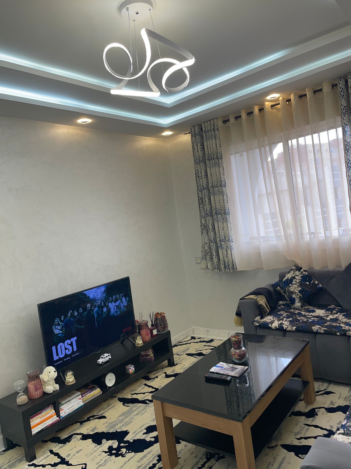A modern living area is bathed in natural light from a large window, adorned with sheer curtains. A dark coffee table sits in front of a comfortable gray sofa, complemented by decorative pillows. A wall-mounted television is set against a smooth white background, enhancing the room's contemporary design.