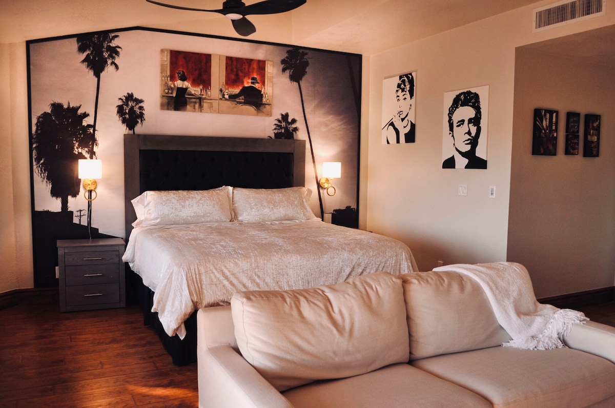 A spacious bedroom is highlighted by a large bed with a light-colored cover. Framed art pieces adorn the walls, including a striking mural of palm trees. A cream-colored sofa is positioned nearby, with a soft throw draped over its arm.