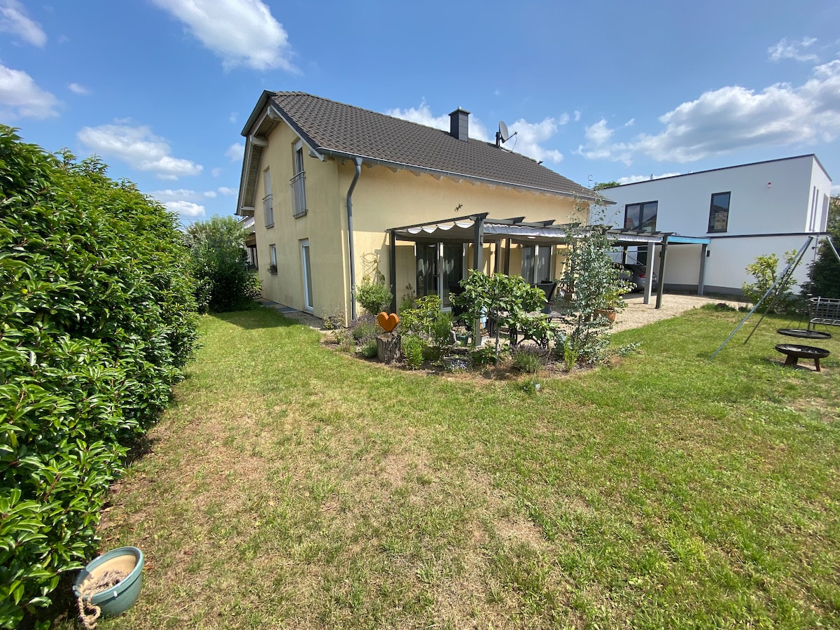 A charming two-story house is surrounded by well-maintained greenery, featuring a covered terrace area with patio furniture. The spacious lawn is complemented by trees and shrubs, creating a serene outdoor environment. Clear blue skies and puffy clouds enhance the peaceful setting.