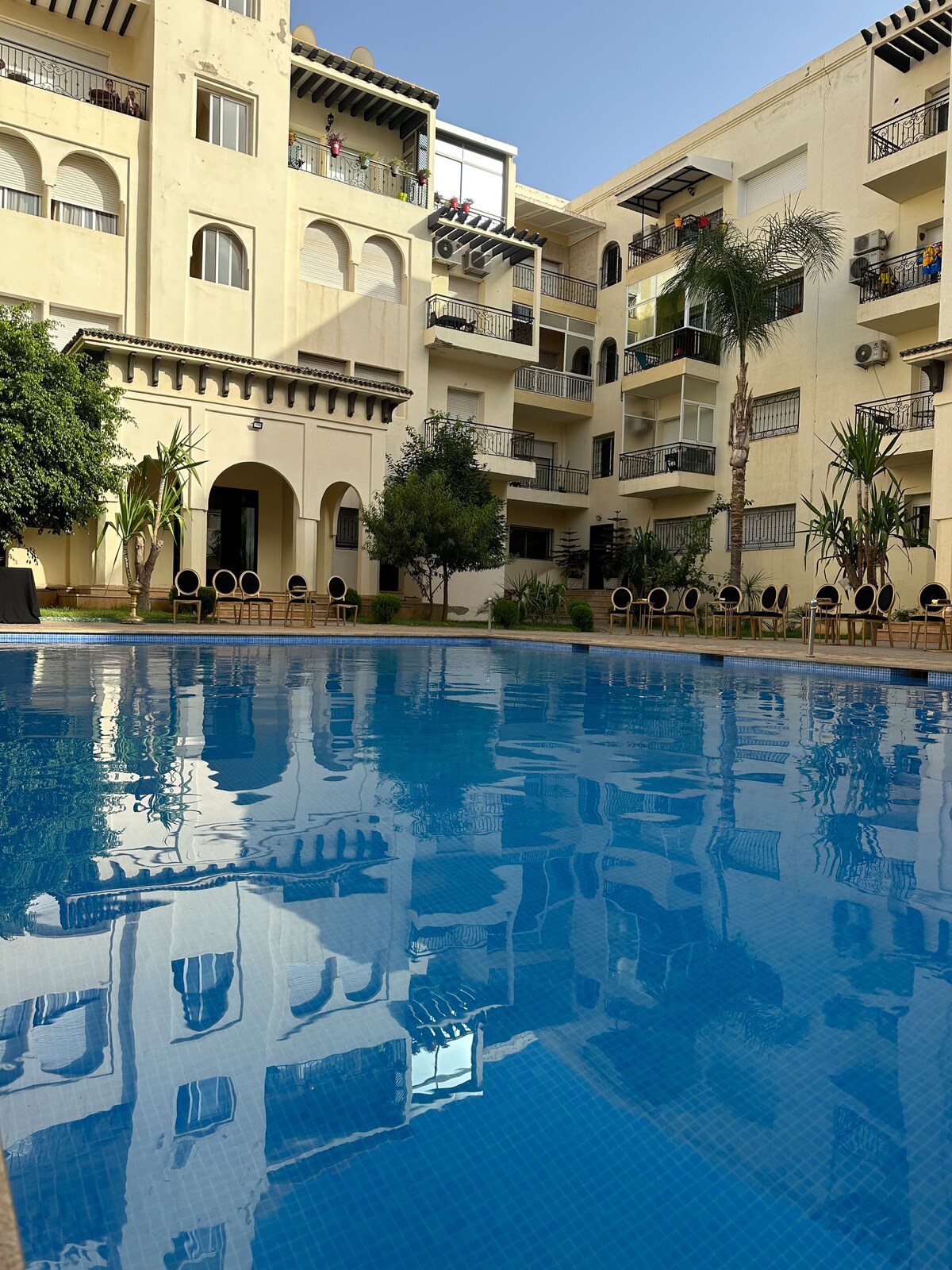 A clear swimming pool reflects the surrounding buildings, featuring balconies adorned with greenery. Lounge chairs are arranged along the pool's edge, with palm trees and well-maintained gardens enhancing the communal outdoor space.