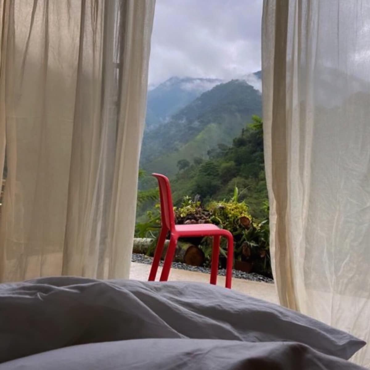 A view from a sleeping area features sheer, flowing curtains framing a red chair placed on an outdoor patio. The backdrop includes lush green mountains shrouded in mist, creating a serene connection with nature.
