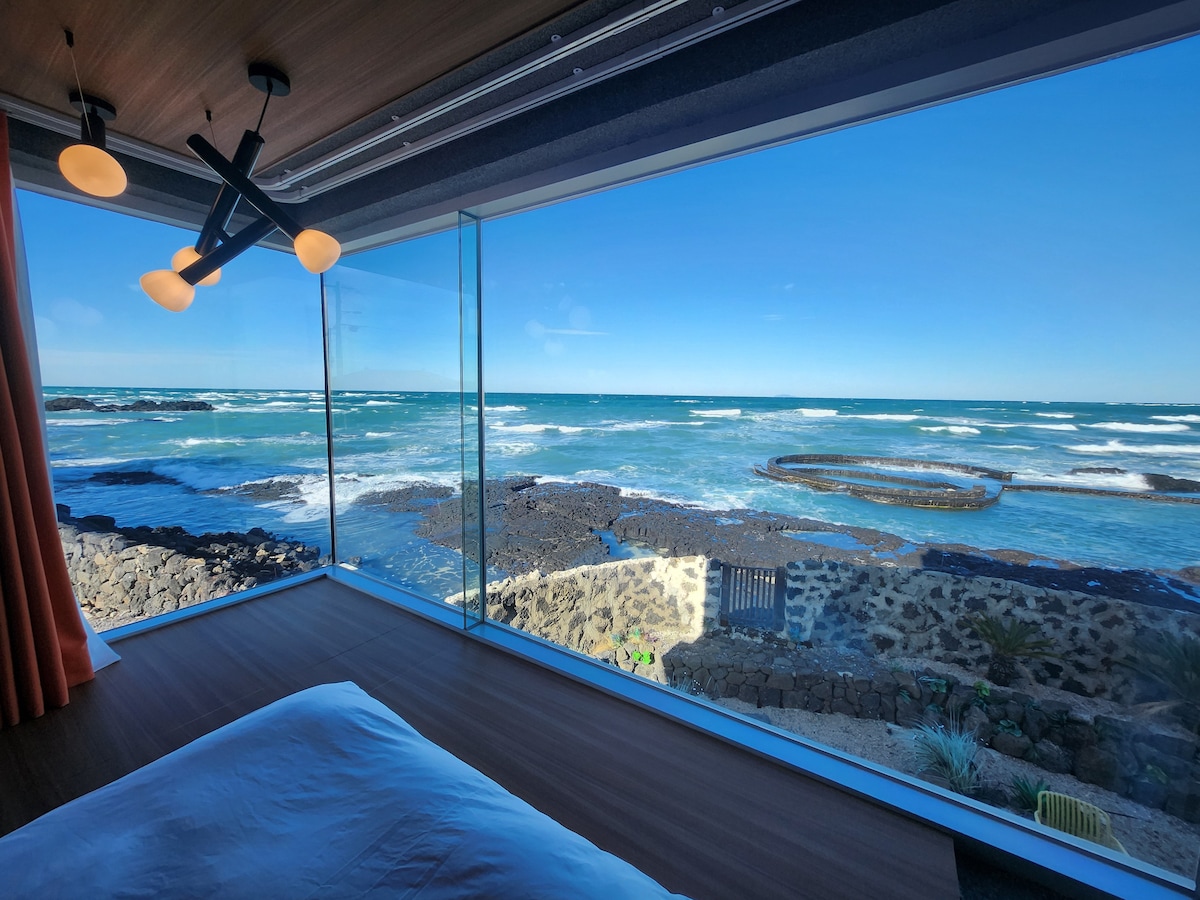 A spacious bedroom features large glass windows providing an unobstructed view of the ocean waves and rocky shore. Natural light fills the room, complemented by modern lighting fixtures and soft drapes. The calming seascape enhances the serene ambiance.