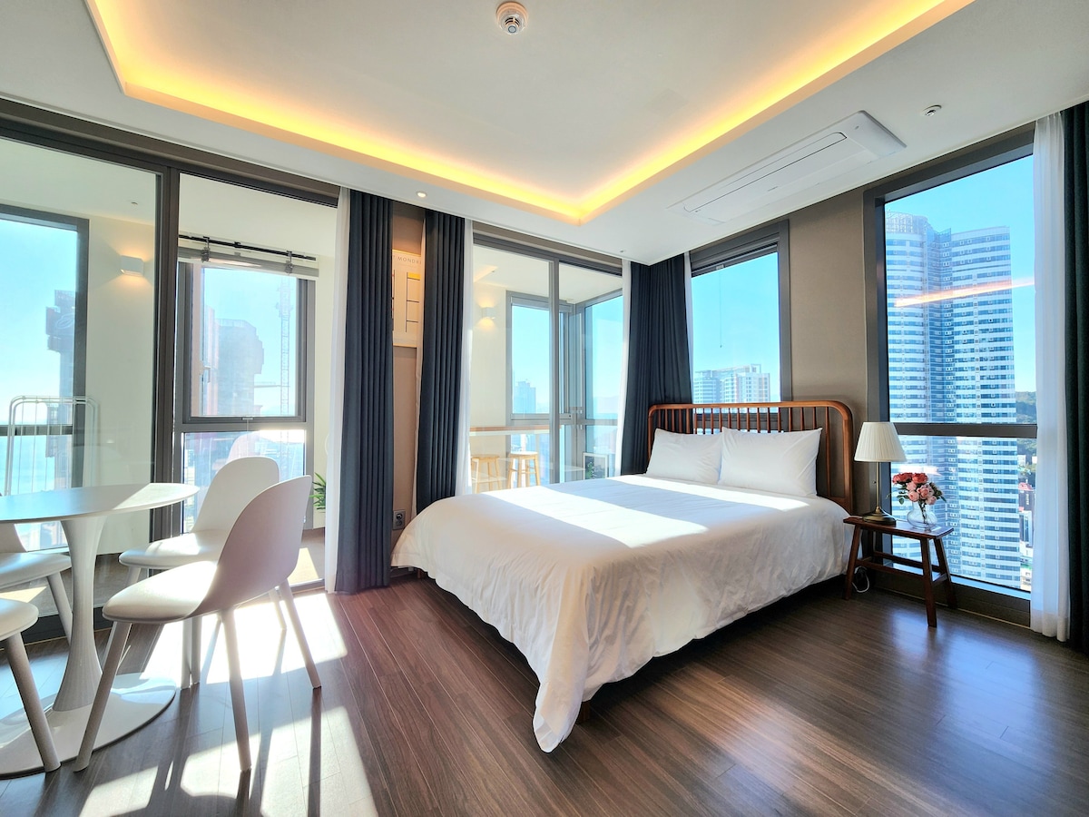 A spacious room features a queen bed with crisp white linens, complemented by large windows offering city views. A round table with four chairs is positioned nearby, and soft lighting highlights the modern decor. Light streams in, enhancing the airy atmosphere.