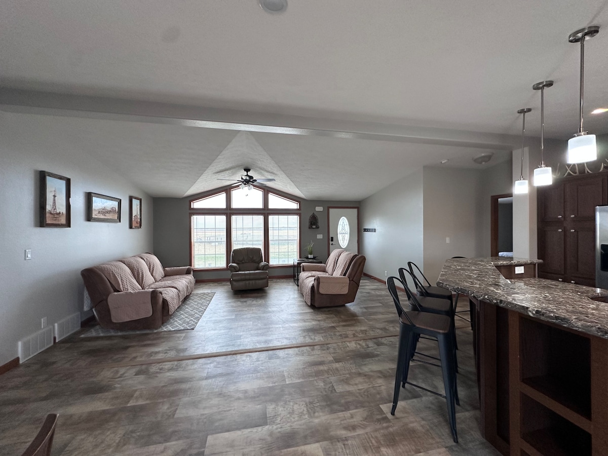 A spacious living area features large windows that provide ample natural light and scenic views of the surroundings. Two comfortable sofas and a recliner create a welcoming seating arrangement, while a modern kitchen island with bar stools complements the open layout.