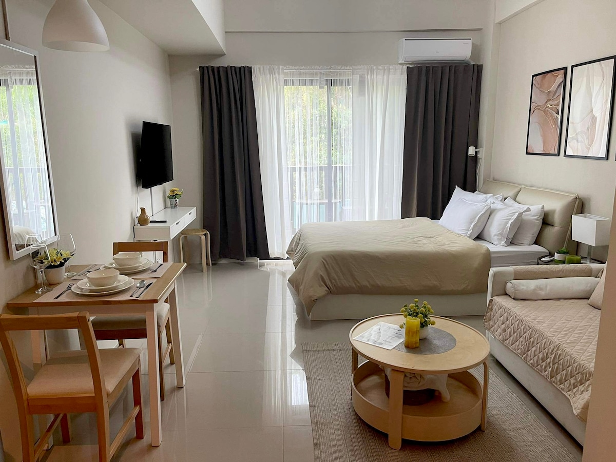 A spacious studio layout is visible, featuring a comfortable bed with neutral bedding and crisp white pillows. A dining area with wooden chairs and a small table is positioned near a large window, with light filtering through sheer curtains. A cozy seating area is included.