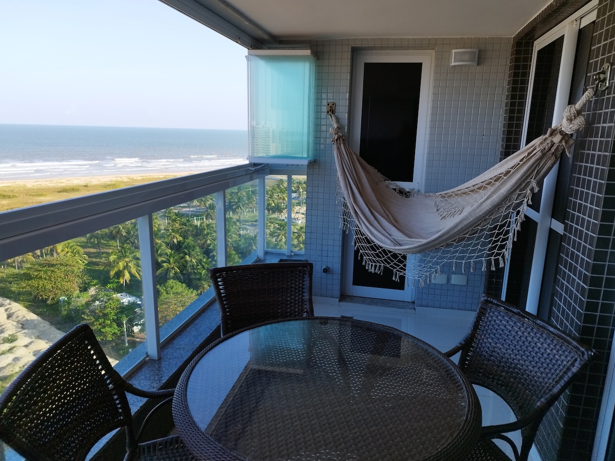 A private balcony offers a tranquil setting with a hammock suspended beside a round dining table surrounded by four wicker chairs. Large sliding glass doors provide access to stunning ocean views, while the gentle ocean breeze is complemented by the serene coastal landscape.