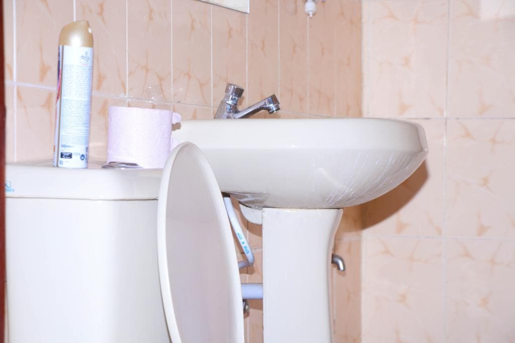 A bathroom area showcases a white sink with modern fixtures positioned next to a toilet. Light pink tiles cover the walls, enhancing the serene ambiance. Toiletry items are placed on the sink, providing organization while maintaining a clean look.