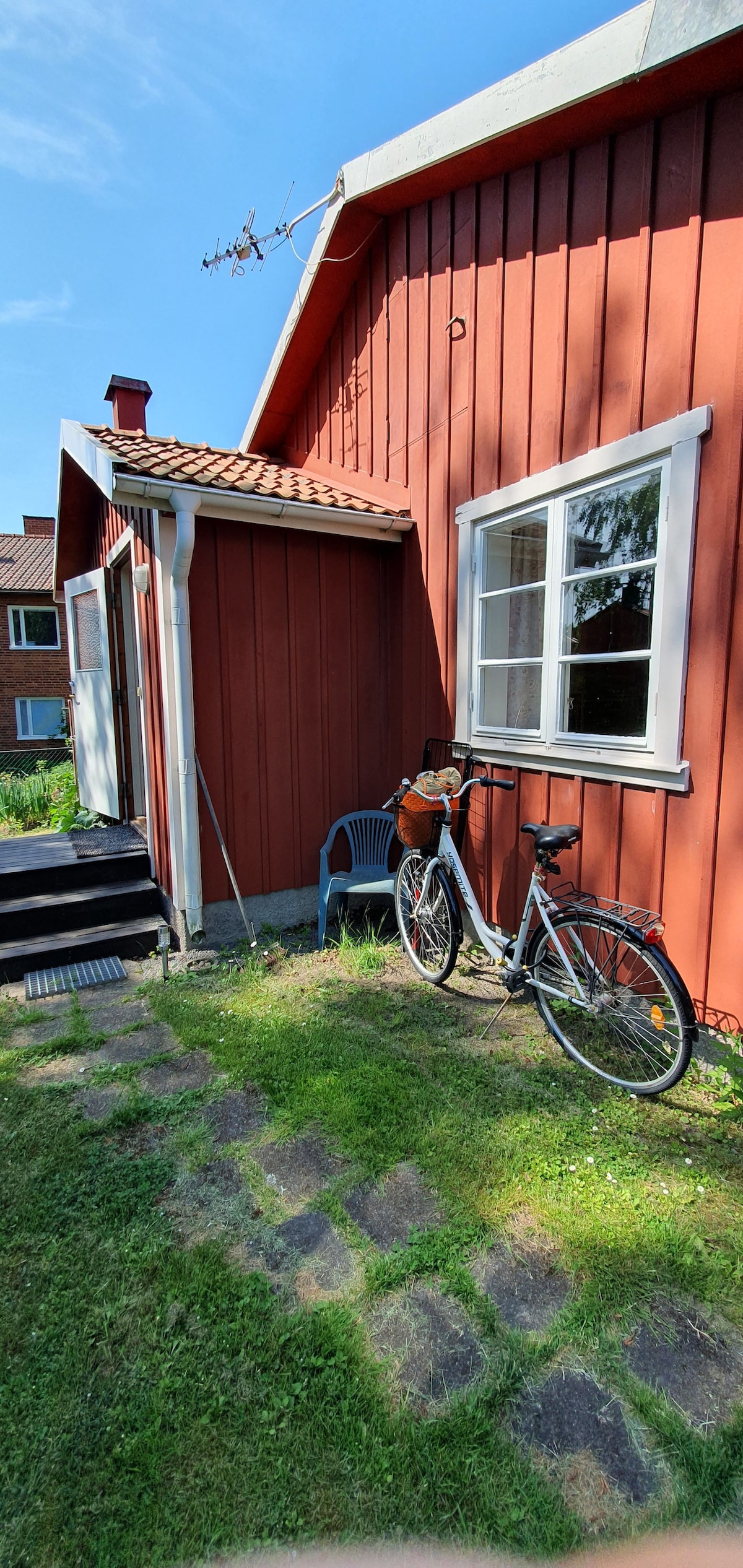 A vibrant red building features a white-framed window and an outdoor area with green grass. A bicycle rests against the exterior wall beside a blue chair, set on a pathway made of stone squares.