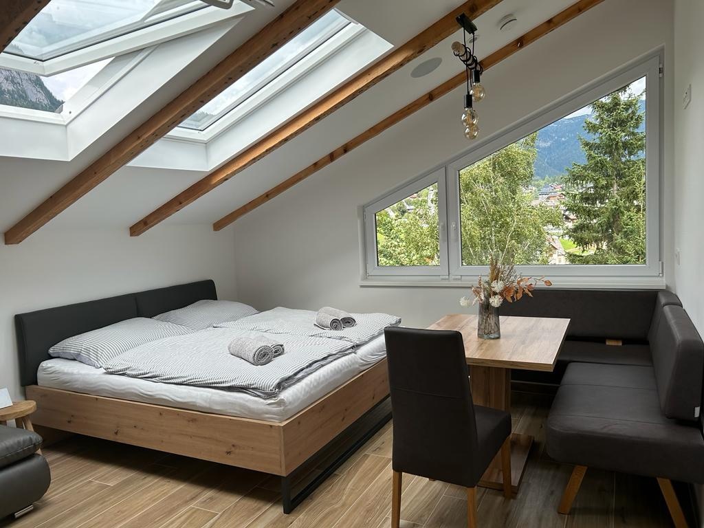 A bright and airy bedroom features two spacious beds with soft linens, complemented by plush towels. A wooden table seats four and is adorned with a simple floral arrangement. Large windows illuminate the room and offer views of the surrounding landscape.