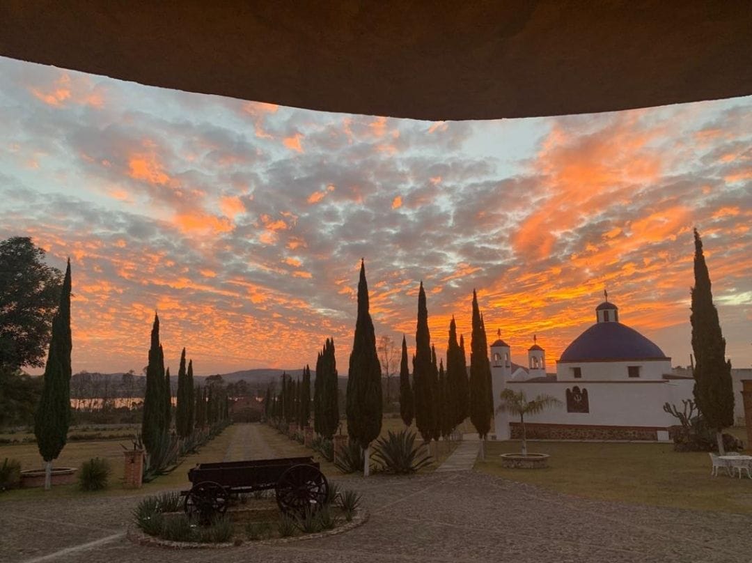 A sunset view captures rolling clouds in shades of orange and pink, framing a pathway lined with tall cypress trees. In the foreground, an antique cart rests on cobblestone, while a charming building with a blue dome and arched windows stands in the background.