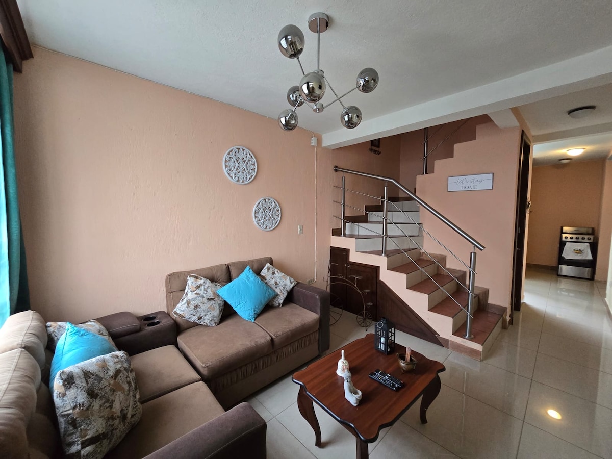 A cozy living area features a brown sectional sofa adorned with decorative cushions and a wooden coffee table. A modern light fixture hangs from the ceiling, while a staircase leads to the upper level. Natural light filters through the window, enhancing the warm peach walls.