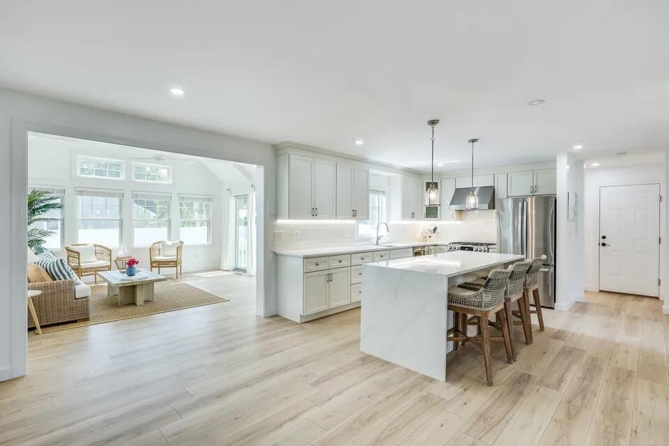 A modern kitchen features light cabinetry, a large island with seating, and stainless steel appliances. Natural light fills the space from adjacent windows, illuminating a connected living area with comfortable seating and decorative accents.