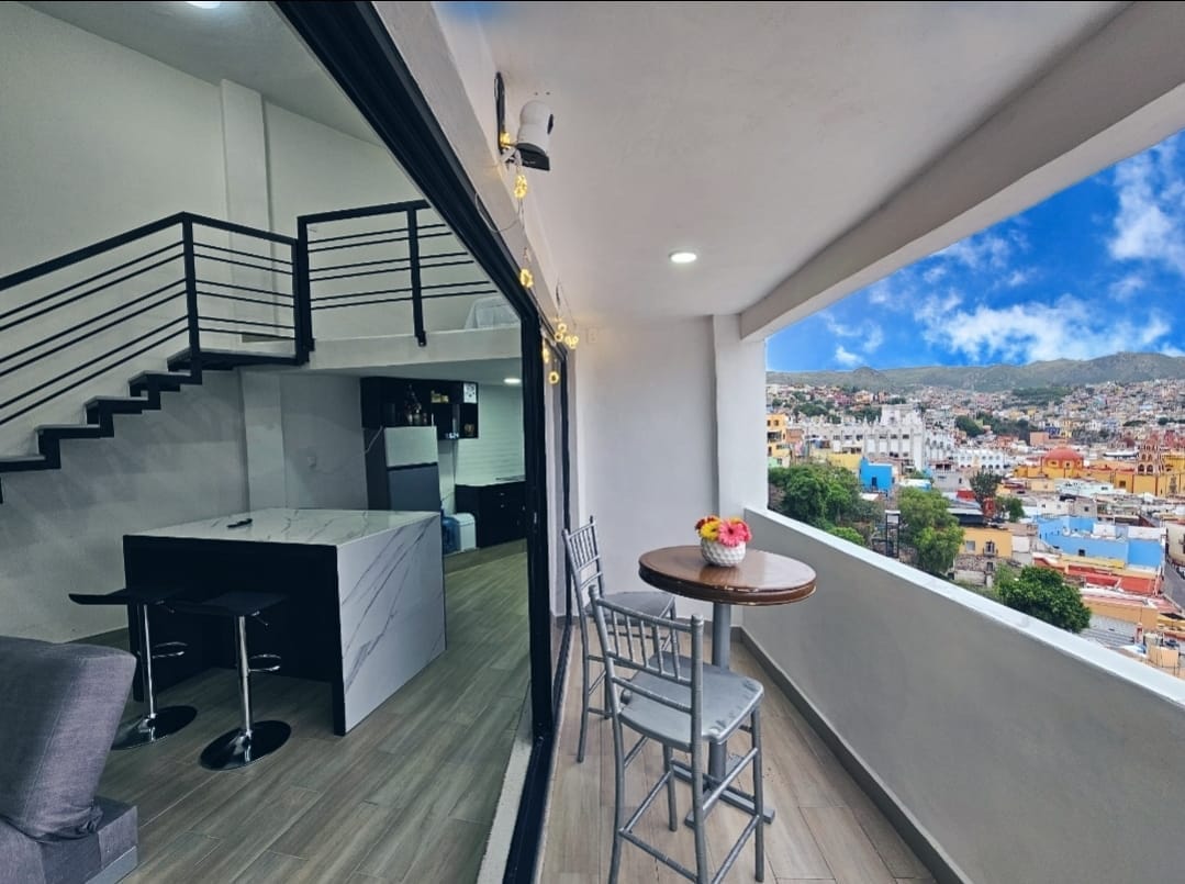 A modern living area features a sleek kitchen and dining space, with a small round table set for two. Large windows frame breathtaking views of Guanajuato, while a staircase leads to an upper level. Natural light brightens the space, creating a welcoming atmosphere.