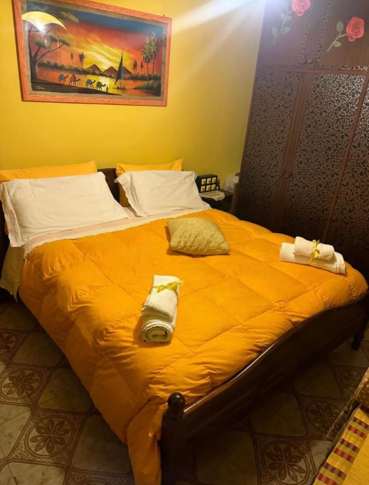 A cozy sleeping area features a spacious bed with yellow bedding and two rolled towels arranged neatly. Decorative elements include a wall art piece, and an intricately designed wooden screen adds texture to the warm yellow walls.