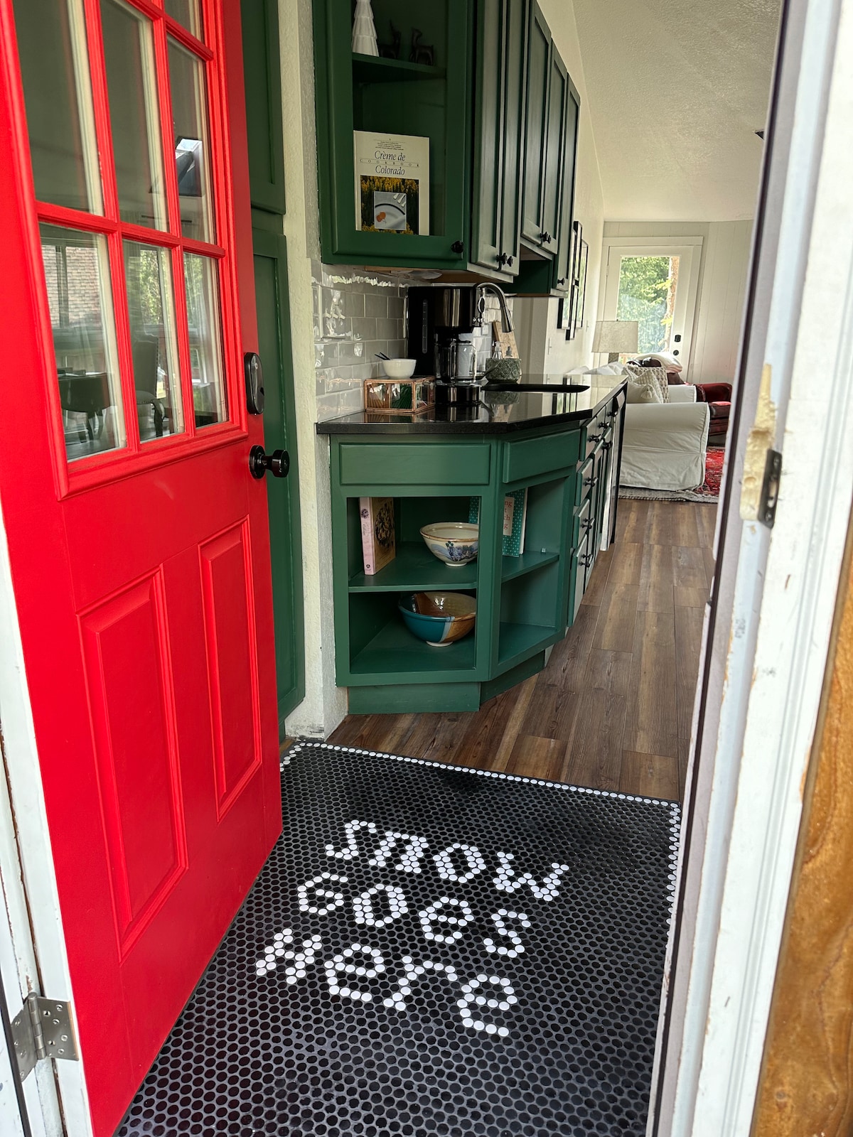 A vibrant red door serves as the entrance to the home, with a welcome mat reading 'SNOW GOES HERE.' Inside, green cabinetry and a coffee station are visible, along with a glimpse of the living area through the doorway.