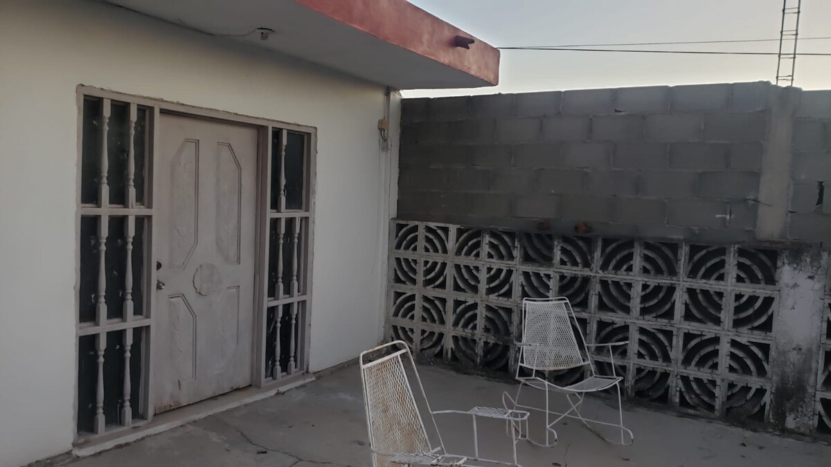 An outdoor area is presented, featuring a solid white door framed by a simple design. Two white metal chairs are positioned on a concrete surface, alongside a textured block wall that enhances the privacy of the space.