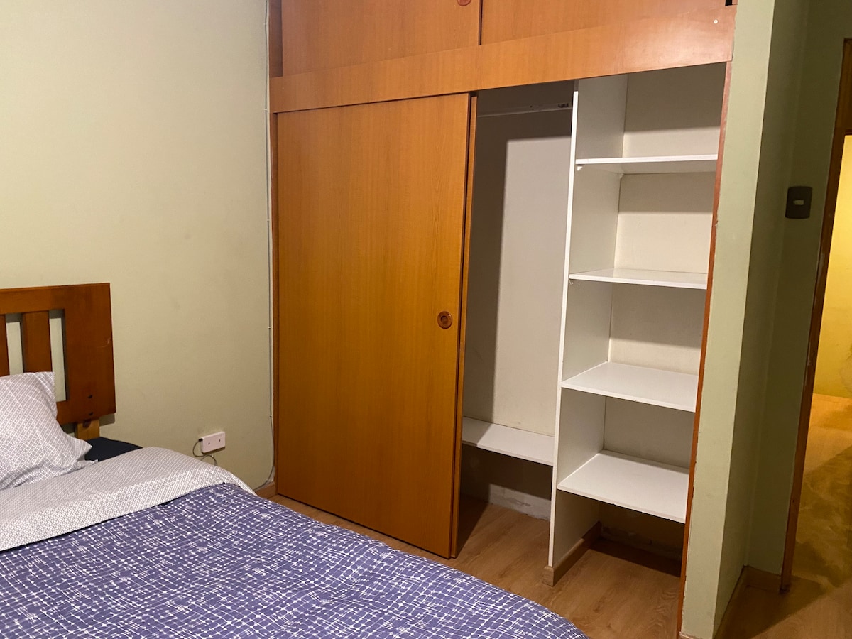 The room features a wooden headboard, a spacious bed with simple bedding, and a built-in wardrobe with sliding doors. Adjacent to the wardrobe, open shelving provides additional storage space. Neutral walls contribute to a calm environment.