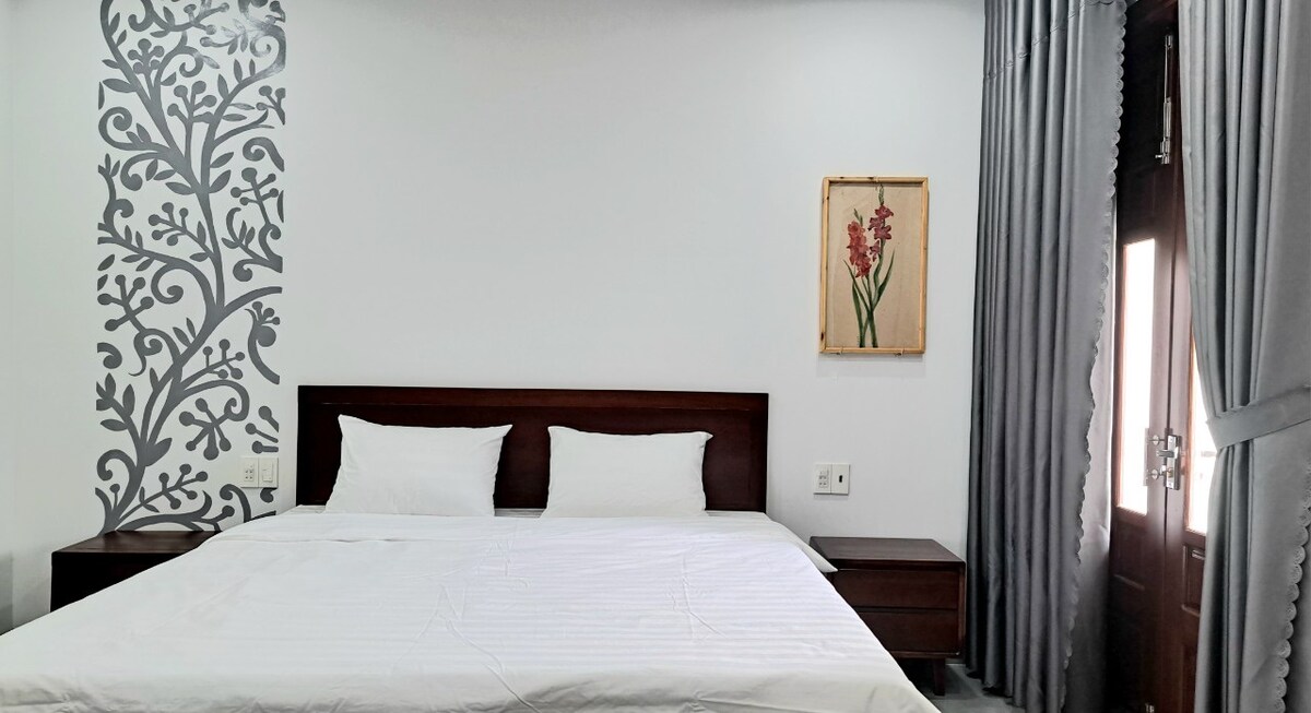 A modern bedroom features a spacious bed dressed in white linens, complemented by wooden nightstands on either side. The wall displays a botanical mural, while curtains frame a window that allows natural light into the room.