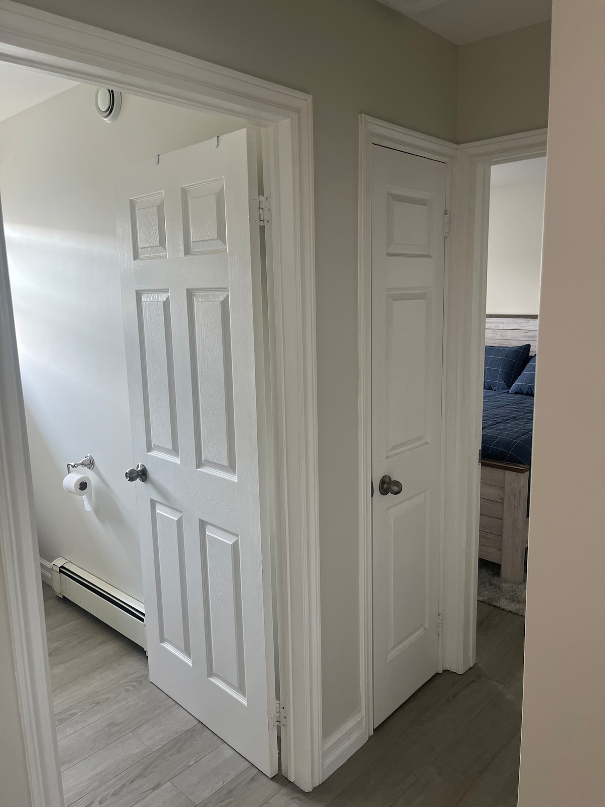 A hallway is depicted, featuring two white doors with chrome handles. One door leads to a bathroom, while the other opens to a bedroom. Natural light enters from a nearby window, illuminating the light-colored walls and wooden flooring.
