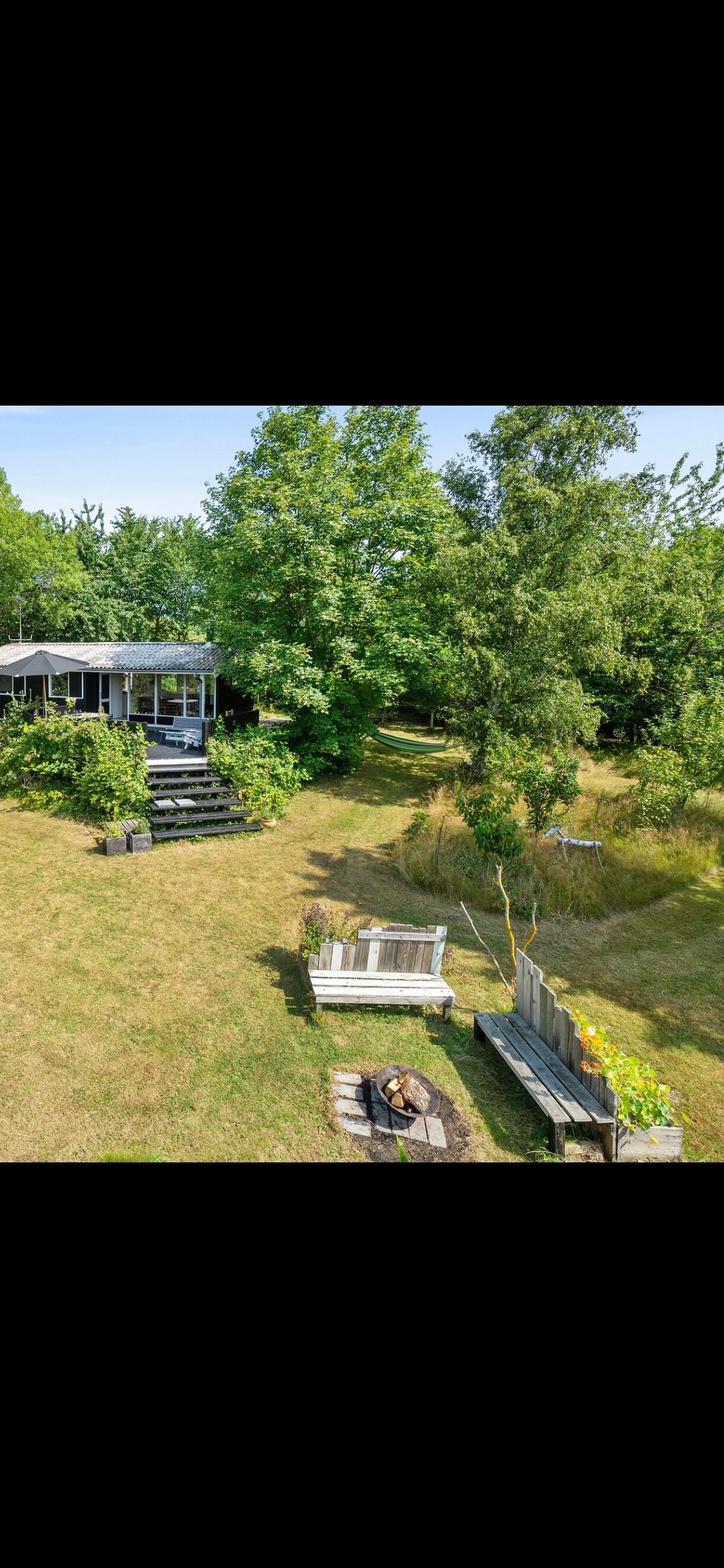 The outdoor area features a spacious lawn bordered by trees, creating a serene atmosphere. A stylish deck is visible with several seating arrangements made from natural materials. A fire pit is surrounded by low benches, providing a cozy focal point for gatherings.