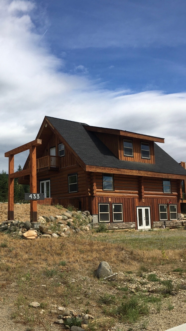 A rustic wooden cabin is displayed with a prominent peaked roof and multiple windows. Natural stone landscaping complements the structure, while an address sign is positioned prominently at the front. The setting is framed by a partly cloudy sky, enhancing the cabin's inviting presence.