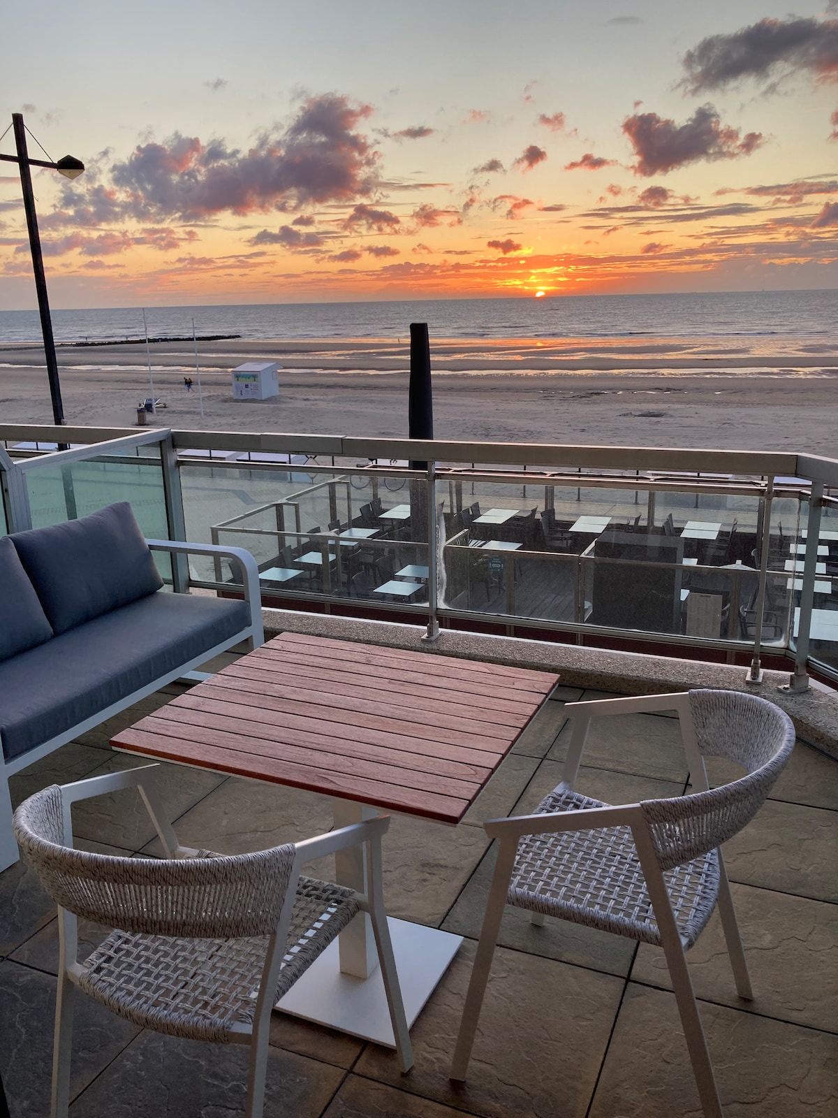 An outdoor terrace features a wood-topped table surrounded by two woven chairs, with a cushioned lounge area visible. The scene captures a sunset over the beach, with vibrant colors in the sky reflecting on the calm water.