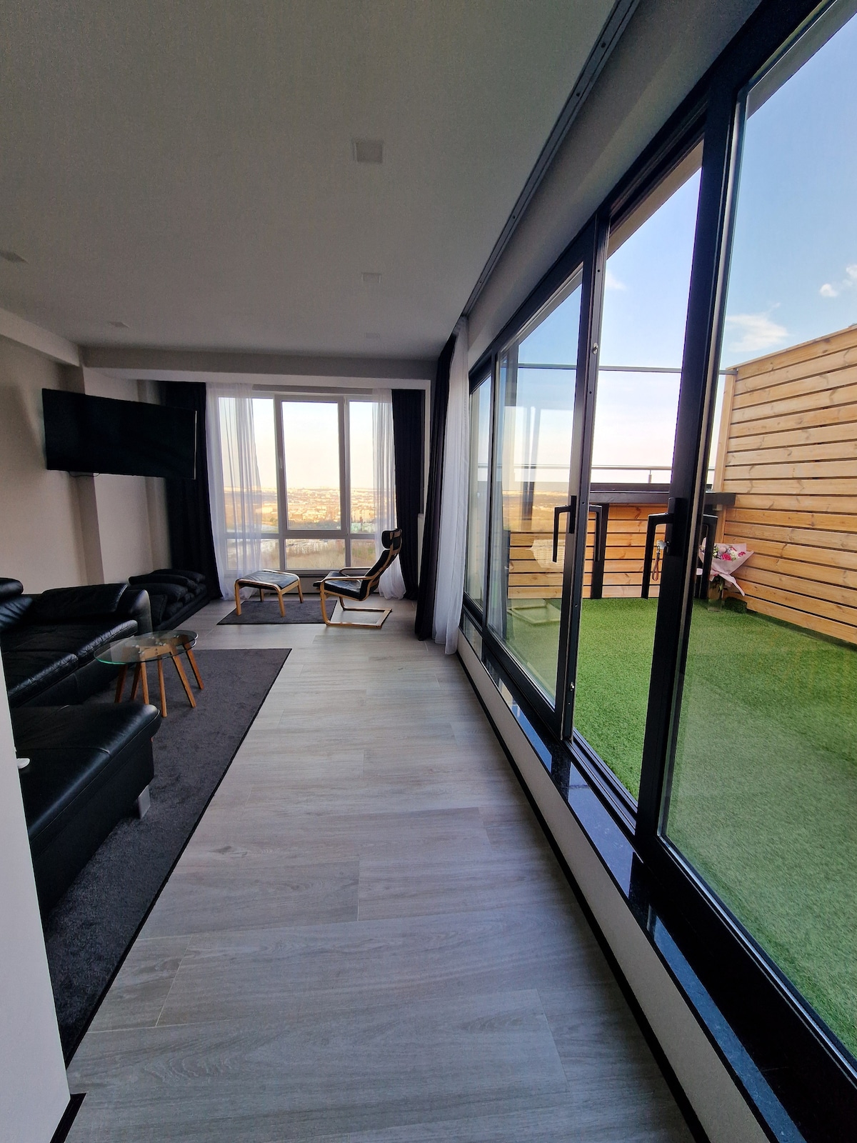 A modern living space features large floor-to-ceiling windows that provide ample natural light. A cozy seating area with black leather sofas is visible, and a television hangs on the wall. The adjoining terrace offers a view of the outdoors and is accessed through sliding glass doors.