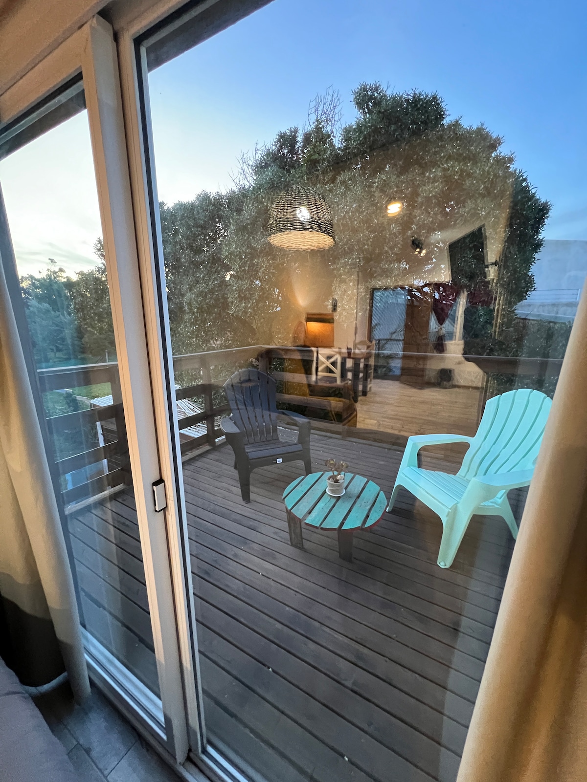 A wooden deck is visible through large glass doors, featuring two chairs and a small table. Lush greenery surrounds the area, providing privacy and a natural backdrop. Soft lighting enhances the relaxed atmosphere, inviting outdoor enjoyment.