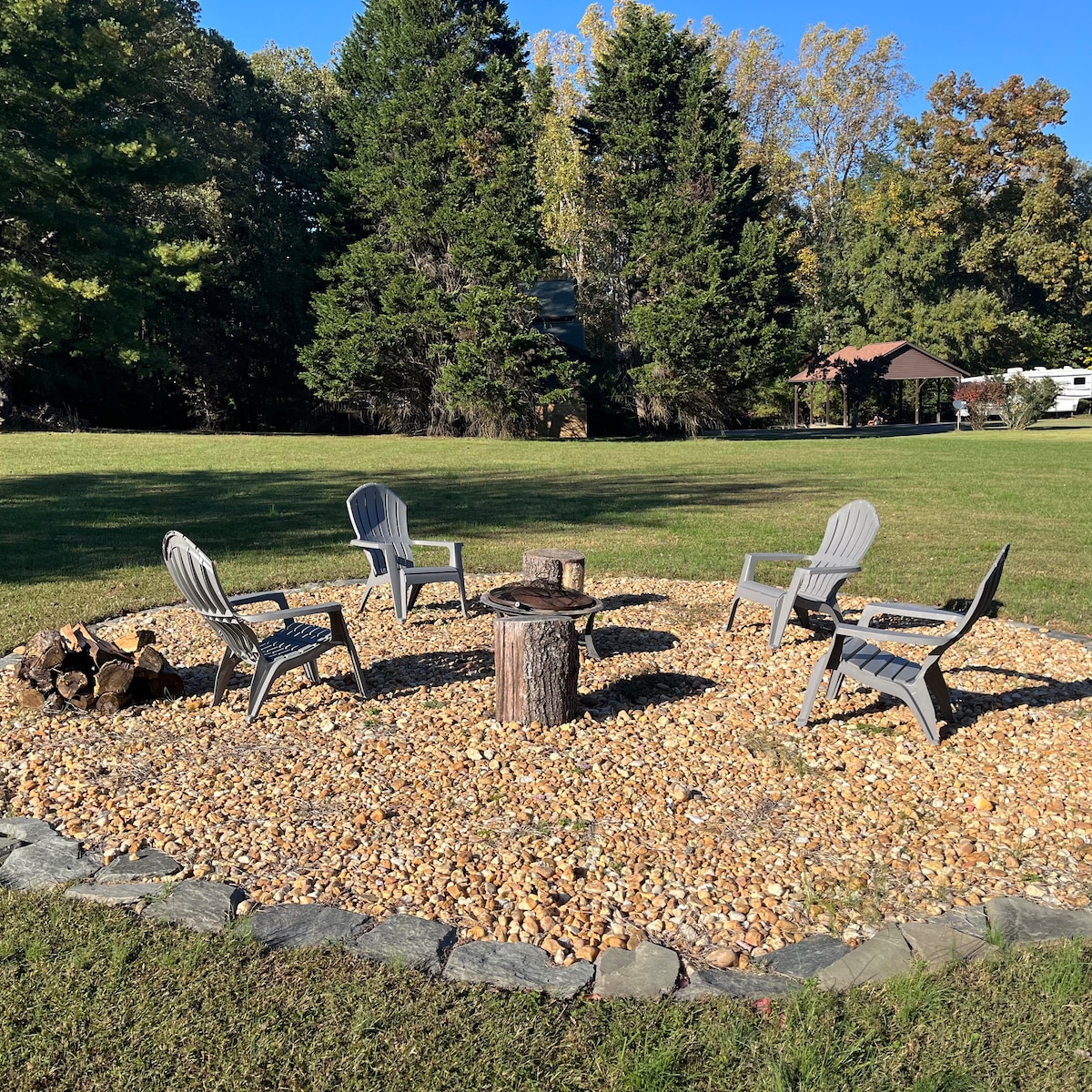 A circular fire pit area is surrounded by smooth pebbles, featuring four grey Adirondack chairs arranged around a central log. Lush greenery and tall trees create a natural backdrop, enhancing the outdoor space's tranquil setting.
