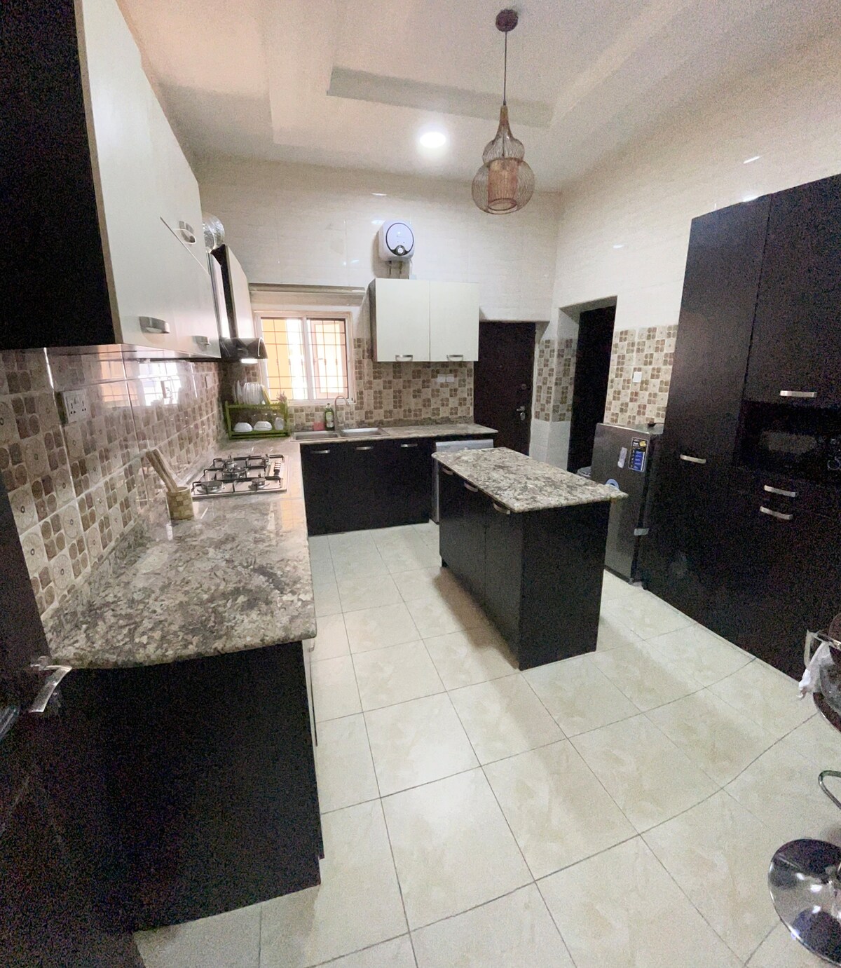 A modern kitchen features dark cabinetry and light countertops, complemented by a tiled backsplash. An island with a smooth surface is central in the space, while various appliances, including a refrigerator and oven, are visible. Natural light enters through a window near the sink.