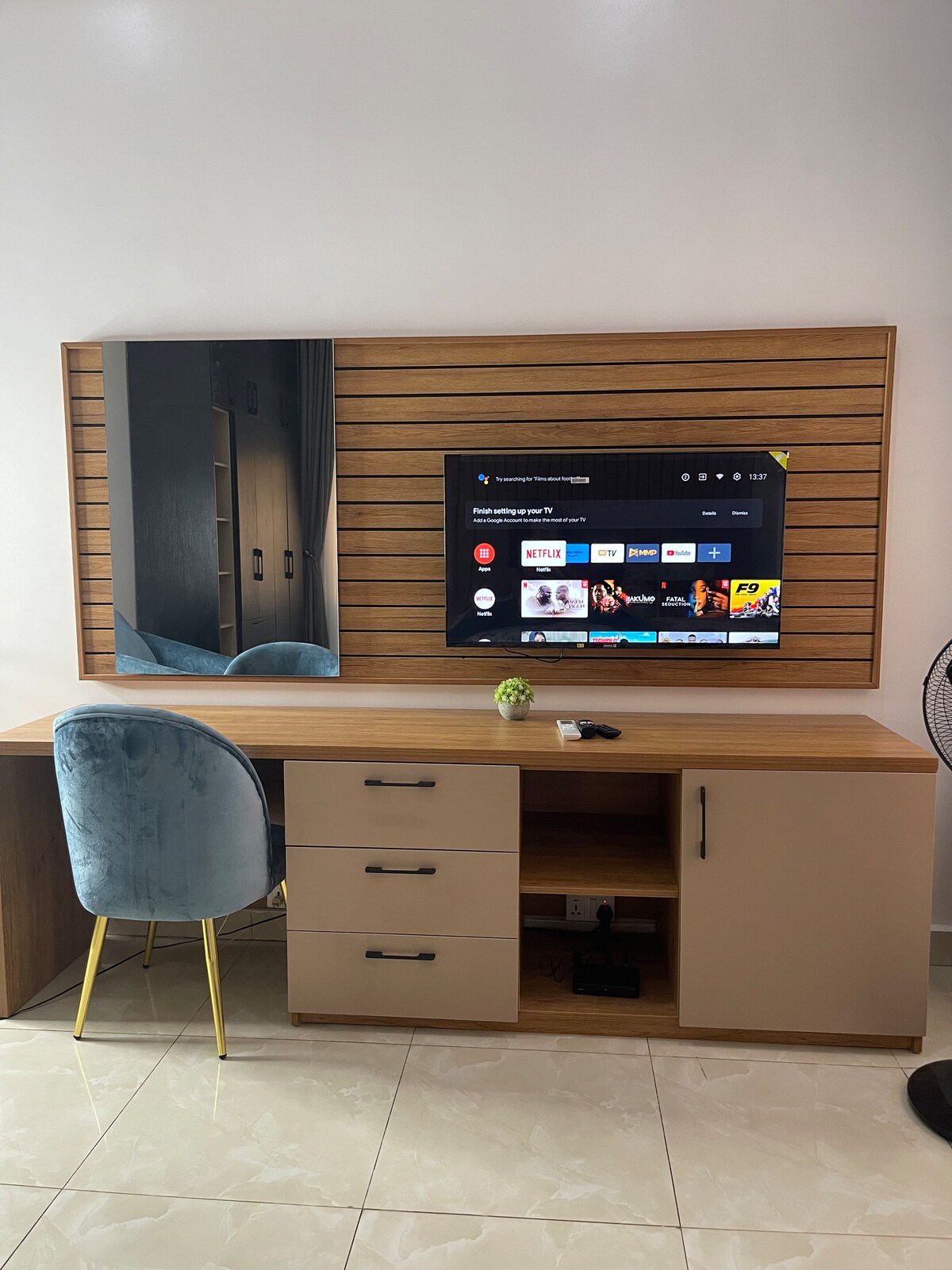 A sleek desk area features a flat-screen TV mounted on a wooden panel, complemented by a large mirror. A stylish blue chair provides seating, while storage cabinets below hold various items. Light-colored tiles enhance the bright atmosphere of the space.