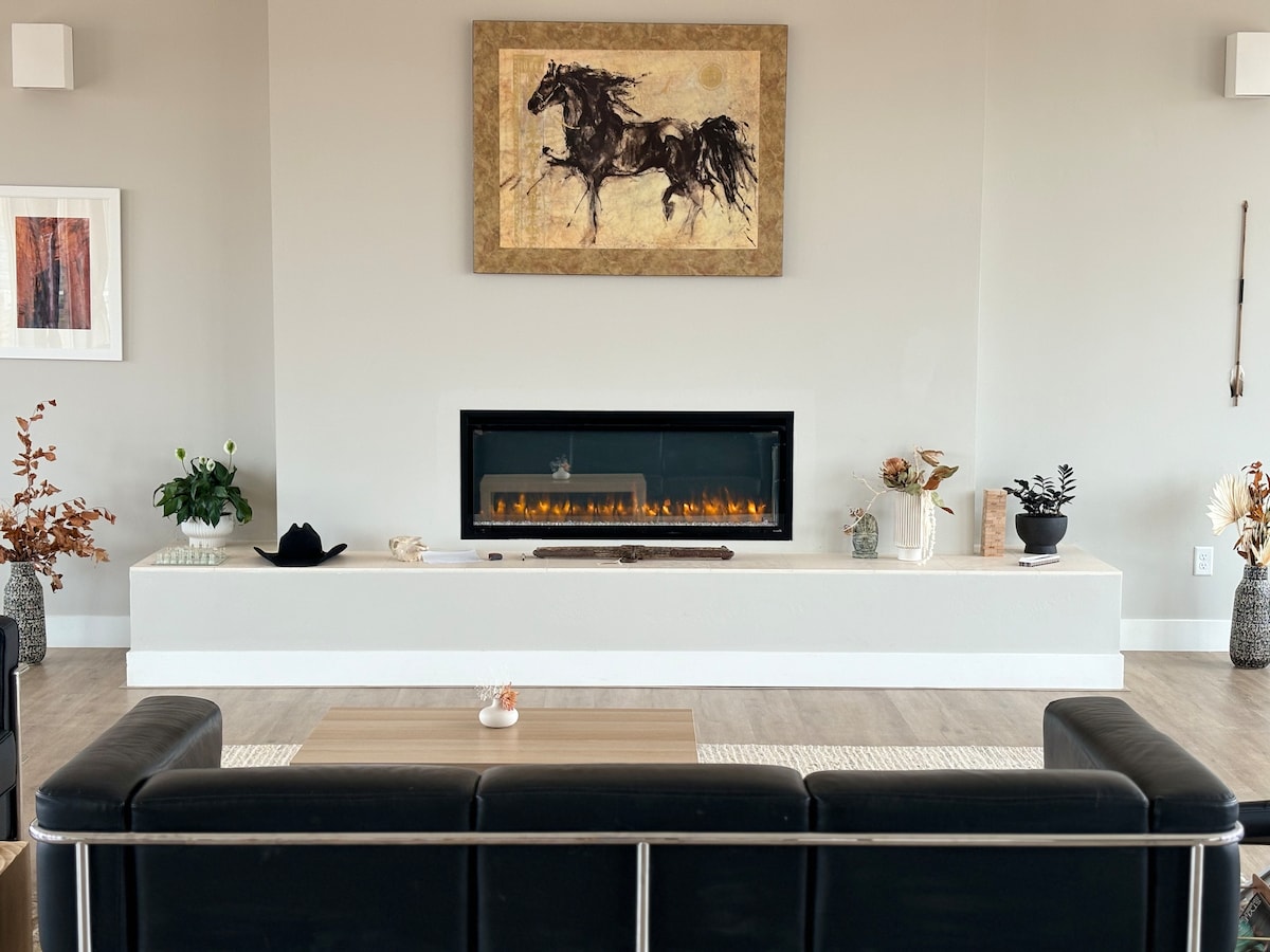 The modern living space features a sleek black sofa positioned across from a minimalist wall fireplace. Above the fireplace, an artwork of galloping horses adds a focal point. Decorative plants and a simple coffee table enhance the ambience, contributing to a clean and open atmosphere.