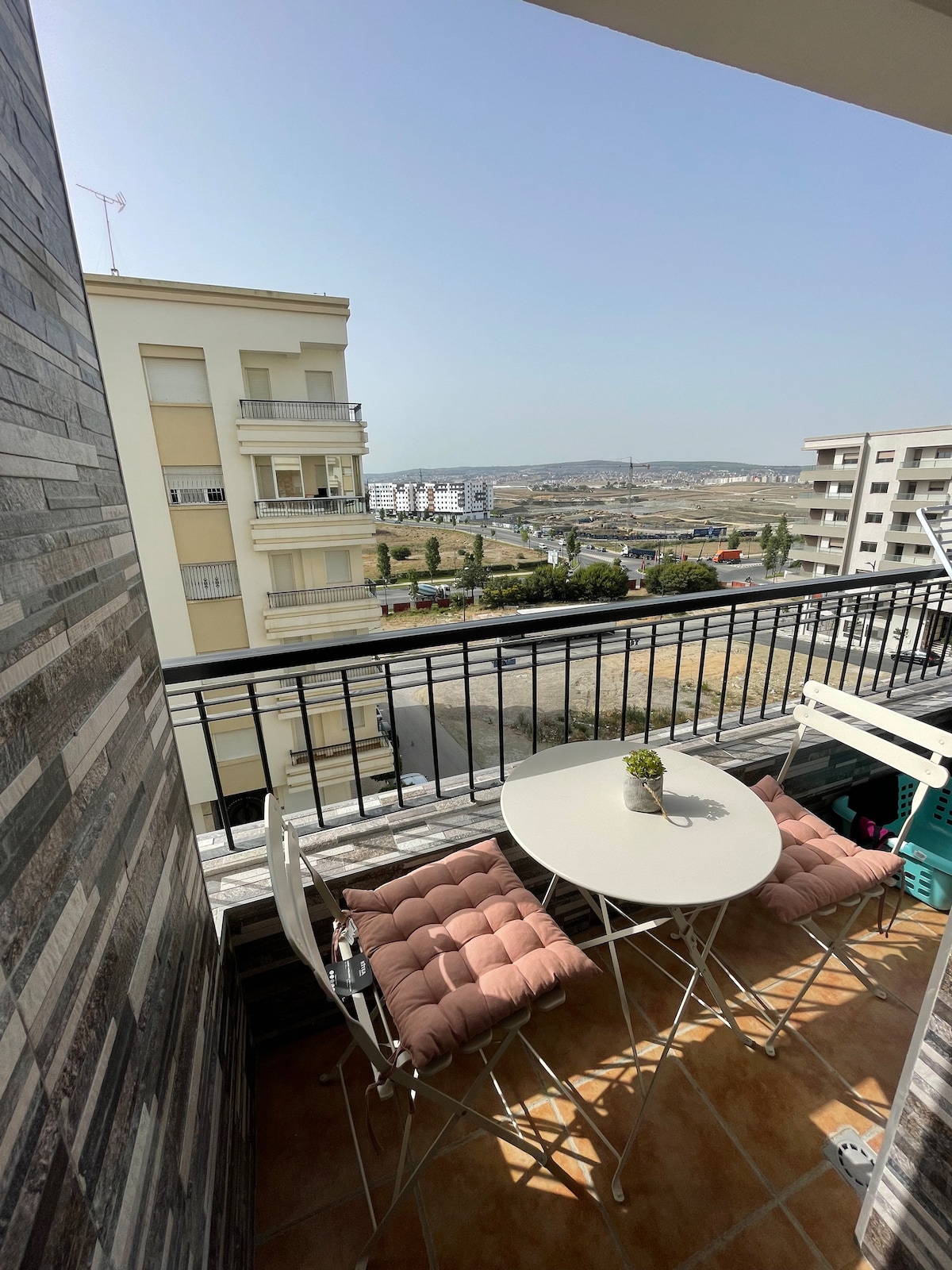 A small balcony is shown, featuring a round table surrounded by two cushioned chairs. A potted plant decorates the table, while views of the surrounding area and distant hills are visible beyond the railing.