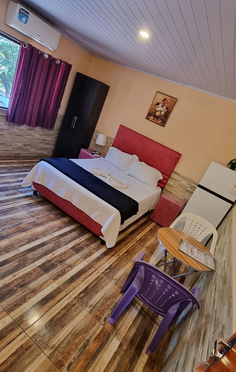 A cozy sleeping area features a spacious bed with a pink headboard and white bedding. A small round table accompanied by two chairs is positioned nearby. A refrigerator stands against the wall, and natural light enters through a window with sheer curtains.